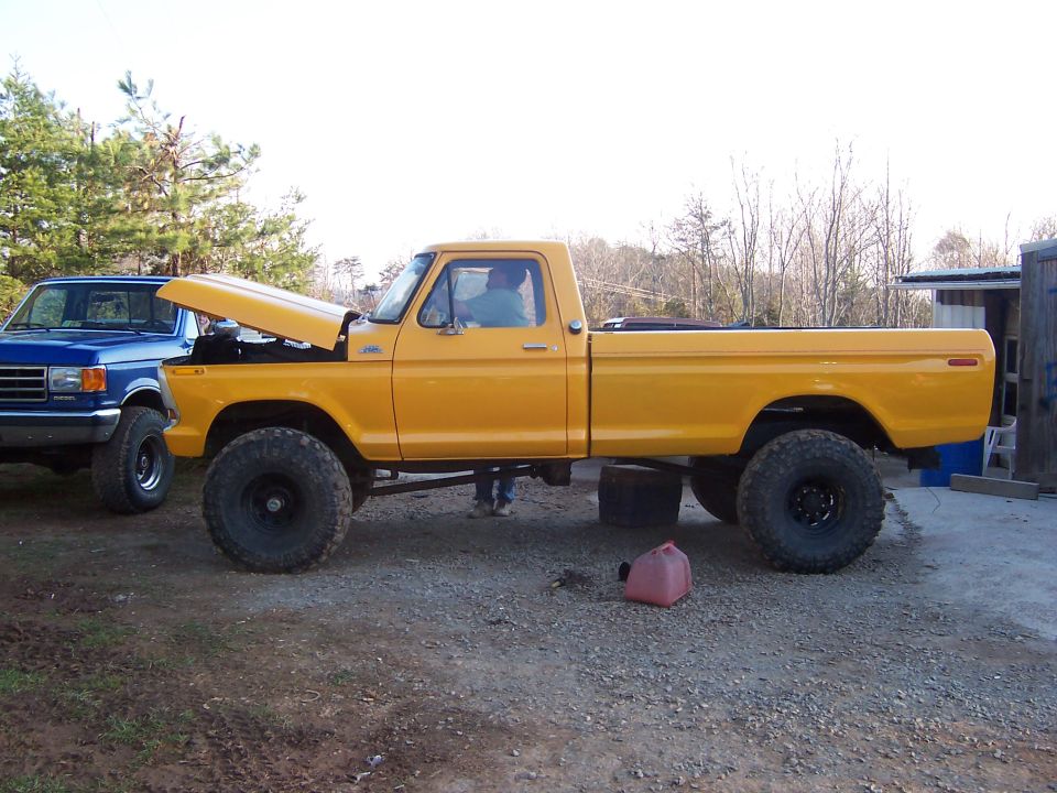 Yellow lifted Ford pickup with hood open beside a blue Ford on gravel, wooded hillside behind
