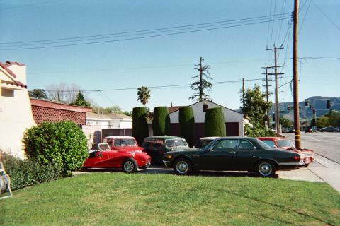 1990 Ford Bronco - Three classic cars including a small red roadster and a dark green coupe parked on a front lawn in a suburban neighborhood.