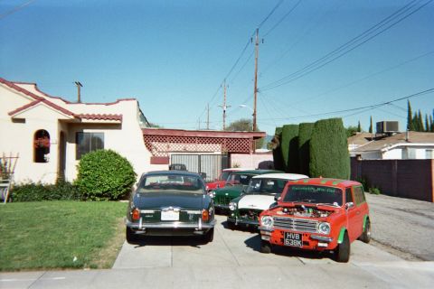 1990 Ford Bronco - Several vintage cars lined up in a driveway, including a red Mini with the hood up and a dark sedan, beside a tiled-roof house.