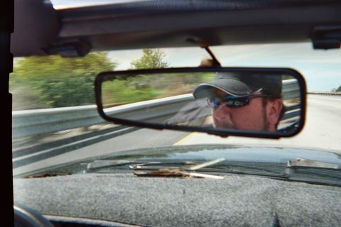1990 Ford Bronco - man wearing a ball cap and sunglasses reflected in a car's rearview mirror while driving along a guardrail-lined highway.