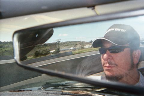 1990 Ford Bronco - bearded man in a dark cap and sunglasses driving, his face framed in the rearview mirror with a hilly highway behind.