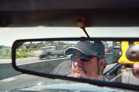 1990 Ford Bronco - mustached man in a cap and sunglasses reflected in the rearview mirror while driving on a freeway with hills in the distance.