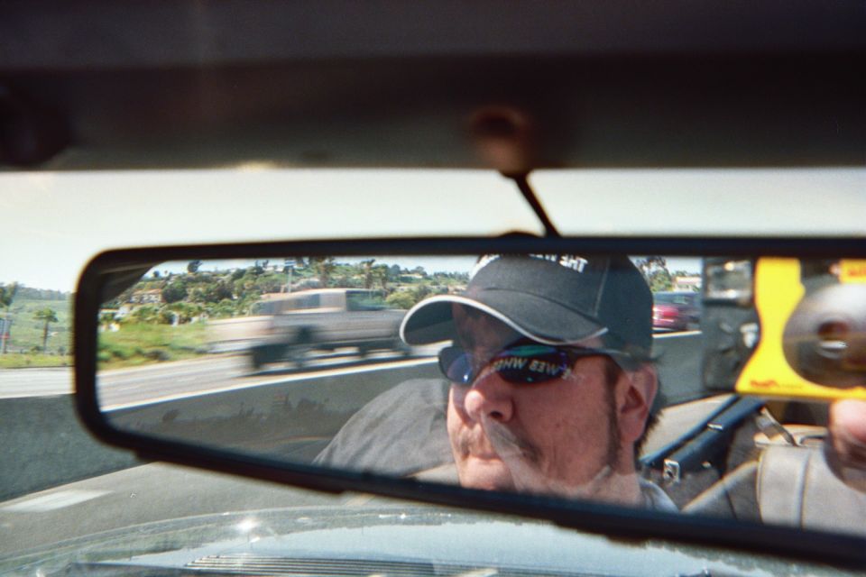 Mustached man in cap and sunglasses driving, reflected in rearview mirror over a freeway
