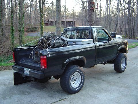 1986 Ford Bronco - A black pickup truck is parked on a concrete driveway, loaded with an engine and secured with ropes. Trees and a house are in the backgroun…