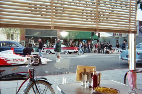 1990 Ford Bronco - View from inside a diner of a downtown street with a white Toyota open-wheel race car and classic cars lined up among pedestrians.