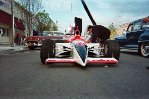 1990 Ford Bronco - white and red open-wheel single-seat race car parked on a town street as a person leans over the cockpit, with classic cars nearby.