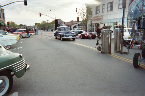 1990 Ford Bronco - small-town street scene with vintage cars including a dark coupe and a green sedan, plus tall gas cylinders on a dolly by the curb.