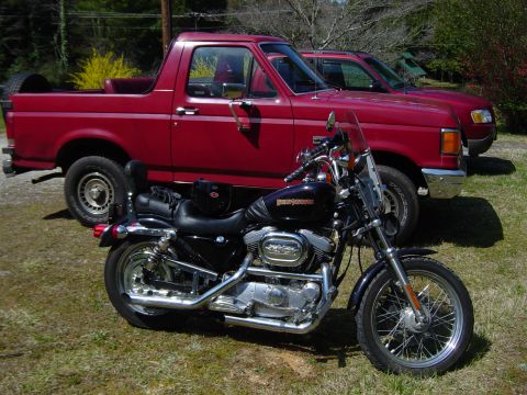 1988 Ford Bronco - A red pickup truck is parked next to a motorcycle, with a second red vehicle visible behind it.