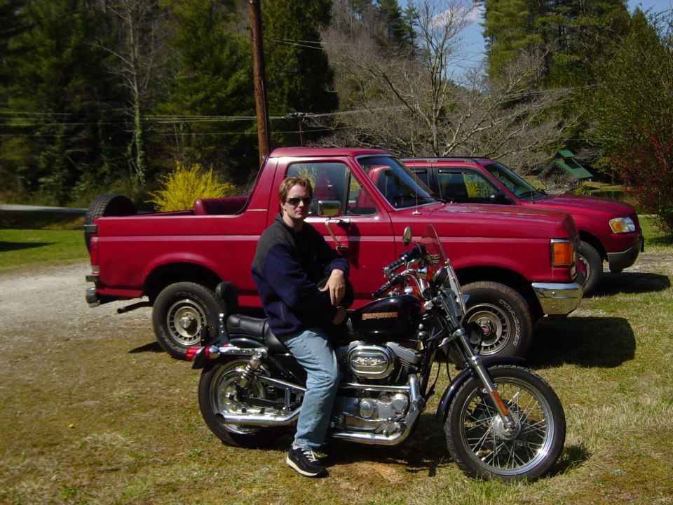 Man sitting on a black motorcycle in front of two red trucks. One truck is a Ford Bronco