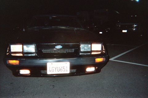 1985 Ford Mustang - A black 1985 Ford Mustang is parked at night, with its front lights illuminated. Another vehicle is parked behind it.