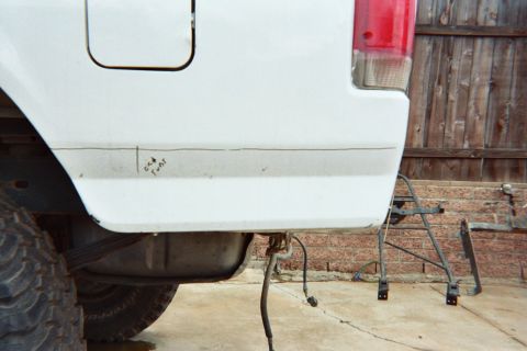 1990 Ford Bronco - Close-up of the rear corner and bedside of a white pickup truck with a red taillight, parked on a concrete pad by a wooden fence.