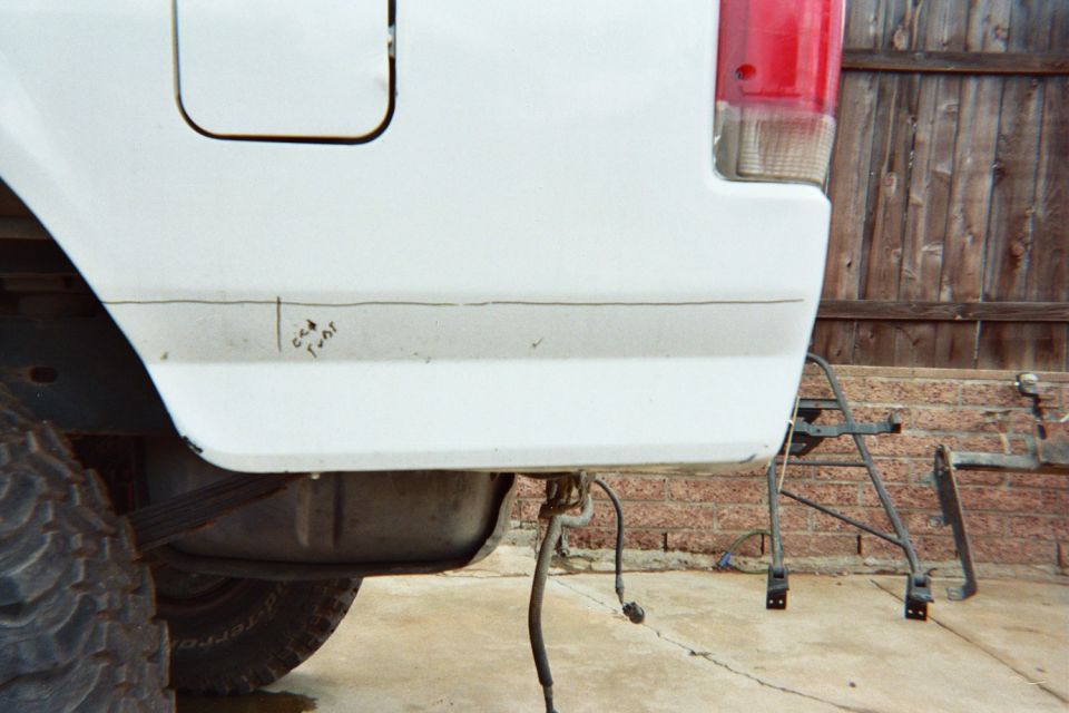 Rear corner of a white pickup truck bedside with red taillight, parked on concrete by a wood fence