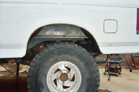 1990 Ford Bronco - rear wheel well of a lifted white truck or SUV showing a chunky off-road tire and chrome alloy wheel on a stained concrete floor.