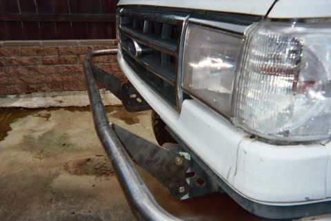 1990 Ford Bronco - Close-up of the front corner of a white Ford truck showing a tubular steel bumper, square headlight and slatted grille against a brick wall.
