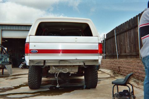 1990 Ford Bronco - Rear view of a lifted white Ford Bronco with a red tailgate stripe and FORD lettering, parked on a wet concrete shop yard by a fence.