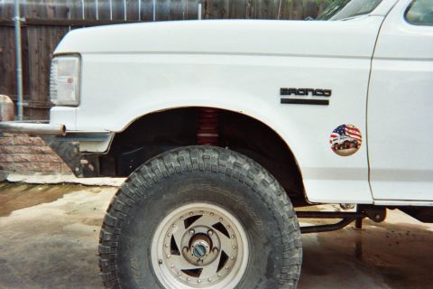 1990 Ford Bronco - Close-up of a white Ford Bronco's front fender with the BRONCO emblem, a USA-flag sticker, an off-road tire and a chrome alloy wheel.