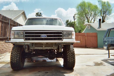 1990 Ford Bronco - Head-on view of a lifted white Ford truck with the oval Ford grille badge and large off-road tires, parked in a fenced backyard.