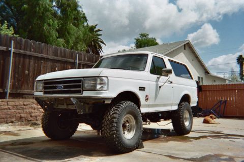 1990 Ford Bronco - lifted white Ford Bronco SUV with large off-road tires and chrome wheels parked on a concrete pad in a fenced suburban backyard.