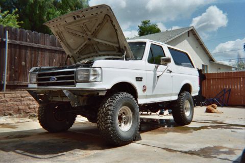 1990 Ford Bronco - lifted white Ford Bronco with its hood propped open, sitting on large off-road tires on a concrete pad in a fenced backyard.