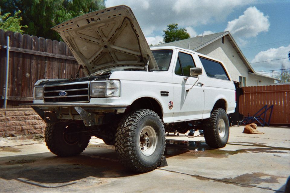 White Ford Bronco with the hood open parked in a backyard, lifted on off-road tires