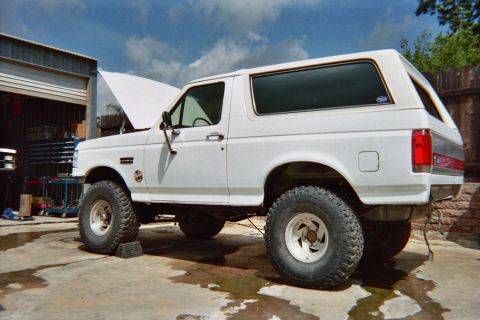 1990 Ford Bronco - Rear three-quarter view of a lifted white Ford Bronco with the hood open, parked on wet concrete next to an open garage.