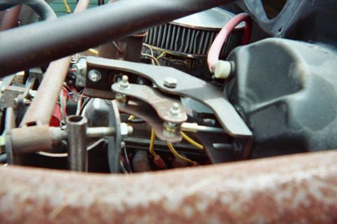 1990 Ford Bronco - Close-up of an engine compartment showing a round air filter, a rusty bolted bracket and linkage with a red wire running through.