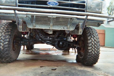 1990 Ford Bronco - View under the front of a lifted Ford truck showing the solid front axle, coil springs and chunky off-road tires on a concrete pad.