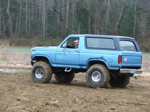 1996 Ford Bronco - A lifted blue Bronco navigates a muddy terrain with trees in the background.
