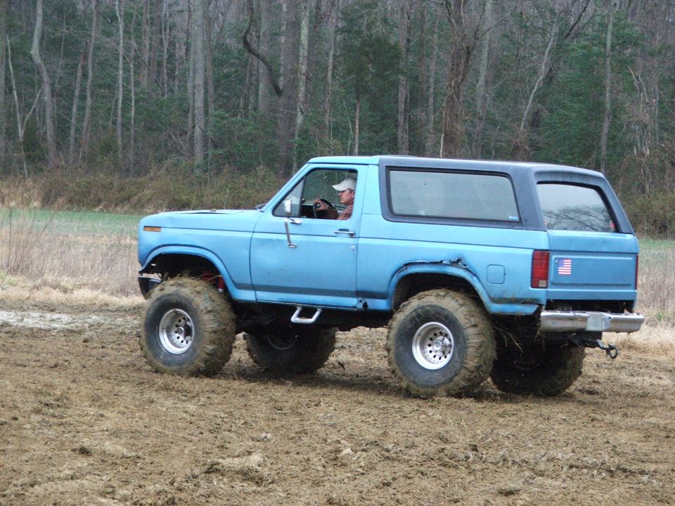 Lifted light blue square-body Ford Bronco on mud tires driving through a muddy field
