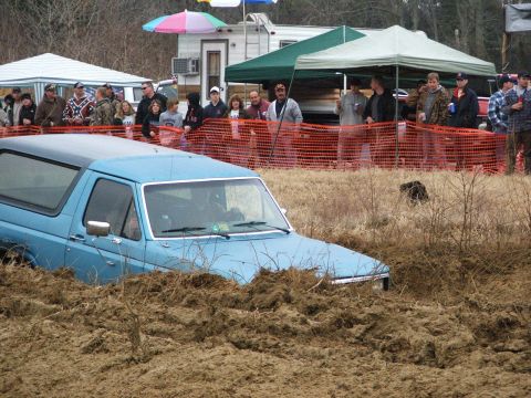 1996 Ford Bronco - A blue 1996 Ford Bronco is partially submerged in thick mud, with onlookers gathered behind a safety fence at a mud bog event.