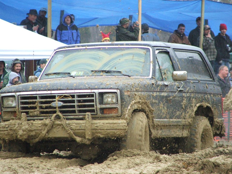 Mud-splattered square-body Ford Bronco churning through bog pit with tow strap on grille