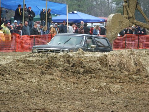 1996 Ford Bronco - A black Ford Bronco is stuck in a large mud pit, with spectators gathered behind safety barriers.