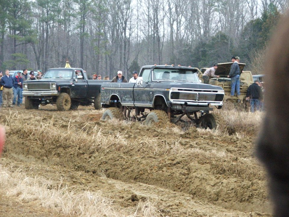 Lifted black Ford pickup on big mud tires stuck in a muddy field at an off-road event with a crowd.