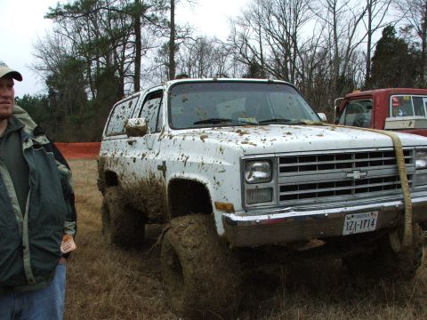 1996 Ford Bronco - A white SUV, heavily coated in mud, sits in a field with a red truck in the background.