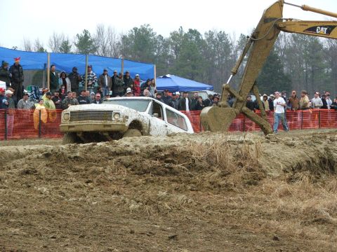 1996 Ford Bronco - A white SUV is stuck in a mud bog, with an excavator arm positioned above it as spectators watch.