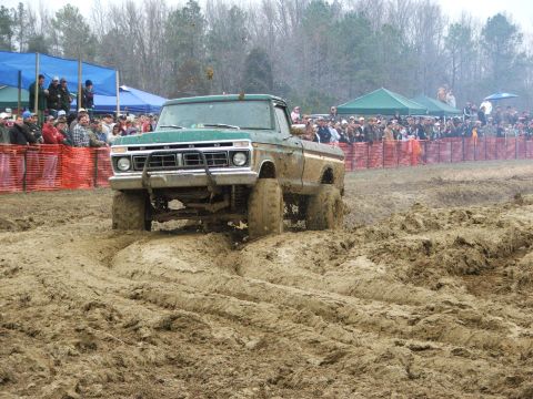 1996 Ford Bronco - A lifted green pickup truck navigates a muddy track during a mud bog event, with spectators watching from behind barriers.