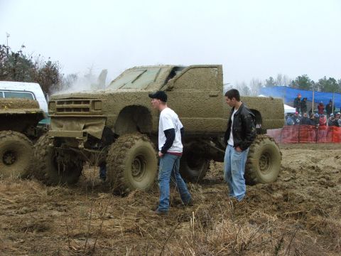 1996 Ford Bronco - Monster truck stuck in mud at an event, with onlookers in the background.