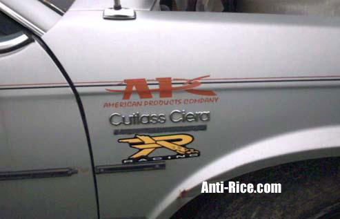 Silver car door with 'AK AMERICAN PRODUCTS COMPANY Cutlass Ciera' and 'X-RACING' decals. Anti-Rice.com and SUPERMOTORS.NET logos visible