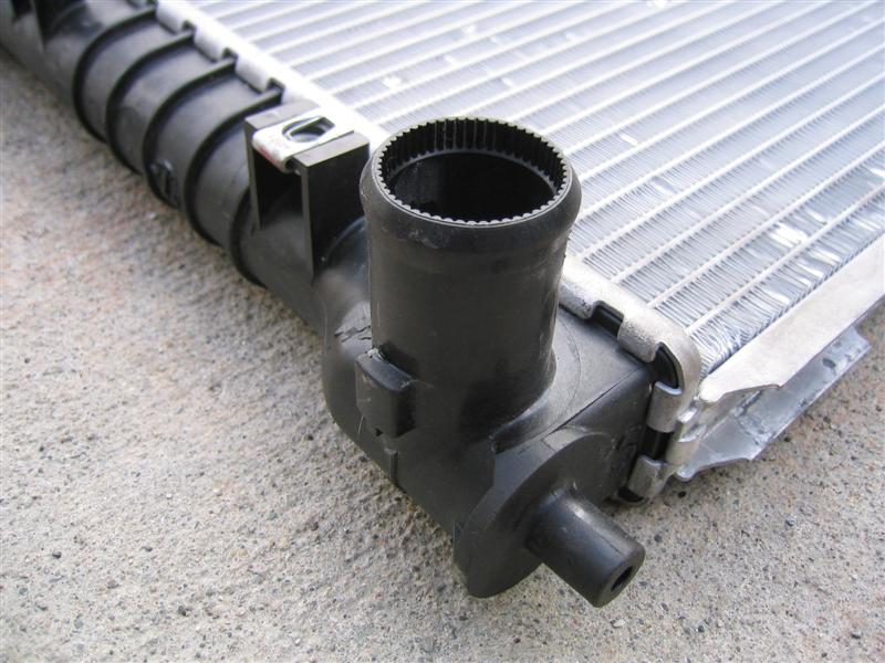 Close-up of a car radiator with black plastic fittings and aluminum fins. Text at bottom says SUPERMOTORS.NET
