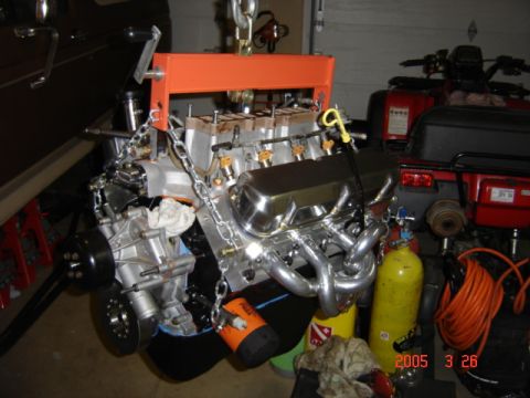 1990 Ford Bronco - Freshly assembled V8 engine hanging from an orange load leveler and chain in a garage, with bare aluminum heads, chrome valve covers, polis…