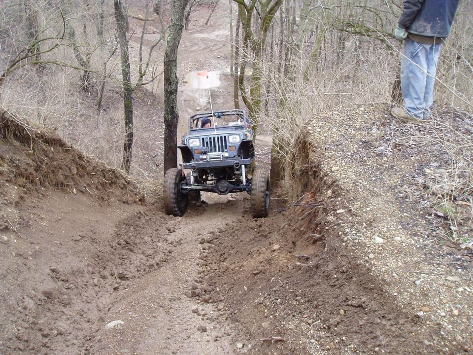 Jeep Wrangler climbing a steep, muddy hill with trees on either side. A person stands on a ridge to the right