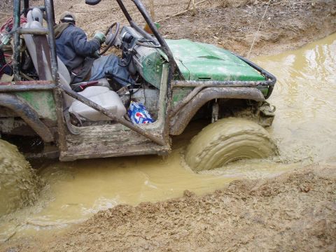 1992 Ford Bronco - A green off-road vehicle with a roll cage is stuck deep in muddy water, with its tires submerged.