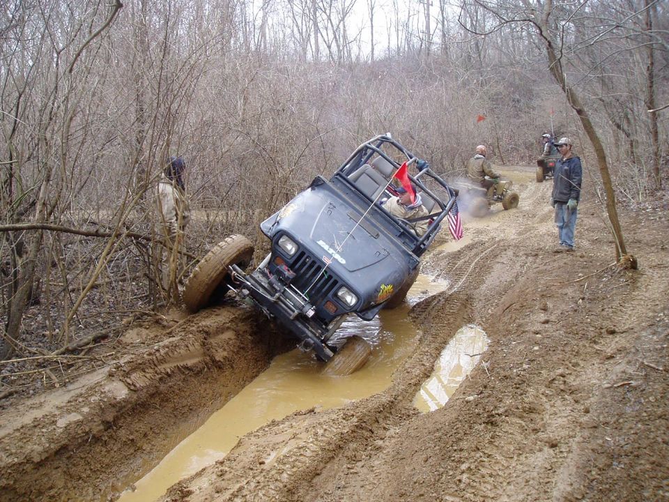 Dark blue Jeep Wrangler tilted in mud pit, with another ATV in background