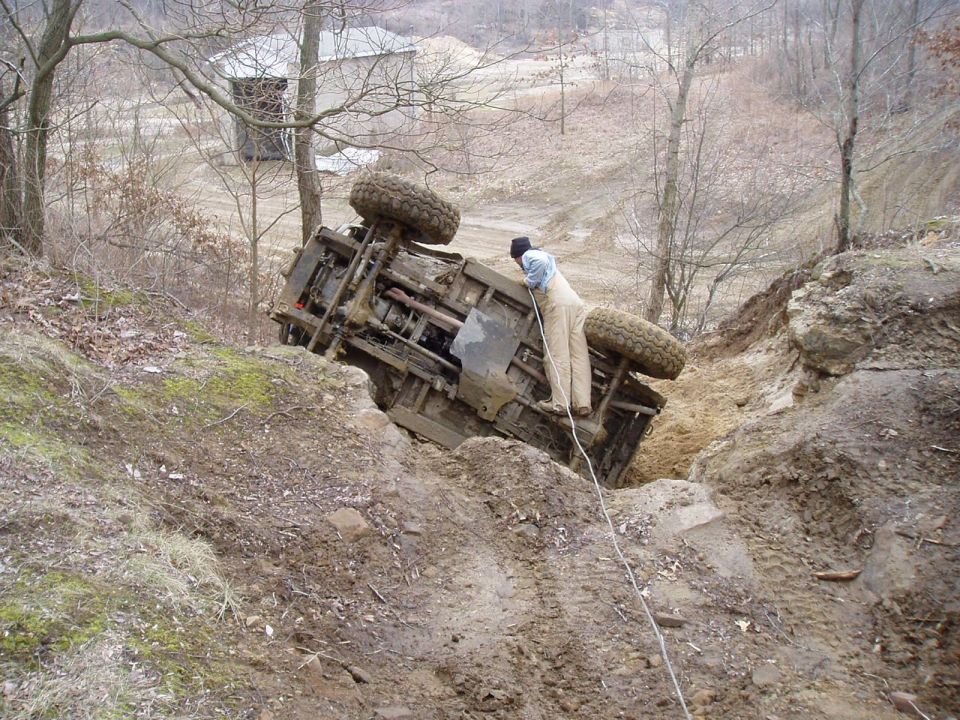 Guy in overalls trying to pull an overturned off-road vehicle out of a ditch with a rope