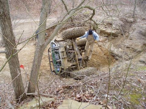 1992 Ford Bronco - A person stands on an overturned off-road vehicle stuck in a ditch, surrounded by trees and dirt.