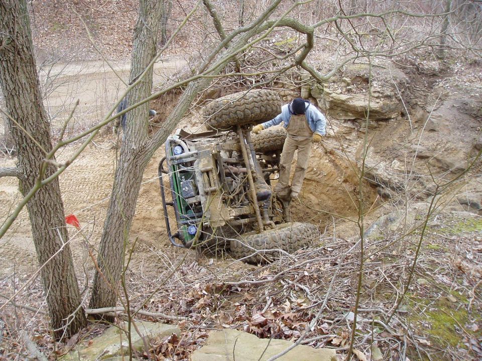 Overturned green off-road vehicle on a dirt embankment with a person standing on its side