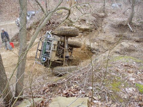 1992 Ford Bronco - An off-road vehicle has overturned on its side in a ditch, with a person observing the scene and another vehicle visible on the road above.
