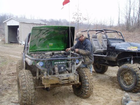 1992 Ford Bronco - Two off-road vehicles are parked on a muddy track, with one having its hood open and a person working on the engine.