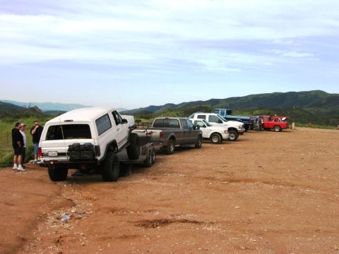 1985 Ford Bronco - Multiple trucks and a white SUV on a trailer are parked in a dusty lot with hills in the background.