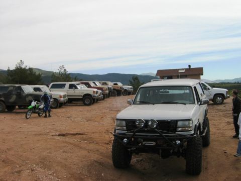 1985 Ford Bronco - A white 1992 Ford Bronco, modified for off-road use, is parked in a lot alongside several other vehicles and a dirt bike.